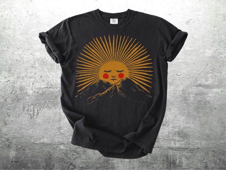 K&ouml;nnte beinhalten: Schwarzes T-Shirt mit einer Grafik einer l&auml;chelnden Sonne mit geschlossenen Augen und rosigen Wangen, die goldene Strahlen &uuml;ber einer Bergkette ausstrahlt. Das Design hat eine Vintage-&Auml;sthetik, handgezeichnet, f&uuml;r den l&auml;ssigen Gebrauch.