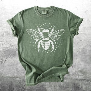 May include: Olive green t-shirt featuring a large white graphic of a bee with outstretched wings and surrounding stars. The tee has short sleeves and a crew neck. The fabric appears soft and slightly faded, giving it a vintage look.