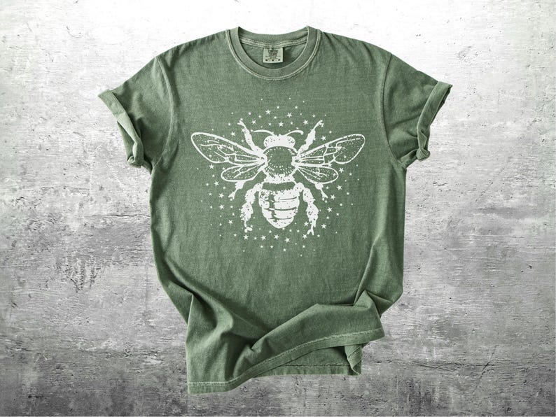 May include: Olive green t-shirt featuring a large white graphic of a bee with outstretched wings and surrounding stars. The tee has short sleeves and a crew neck. The fabric appears soft and slightly faded, giving it a vintage look.