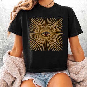 May include: A black short-sleeve t-shirt featuring a graphic print of a central eye surrounded by radiating golden-yellow lines. The eye has a red iris and a black pupil. The design is centered on the chest of the garment.