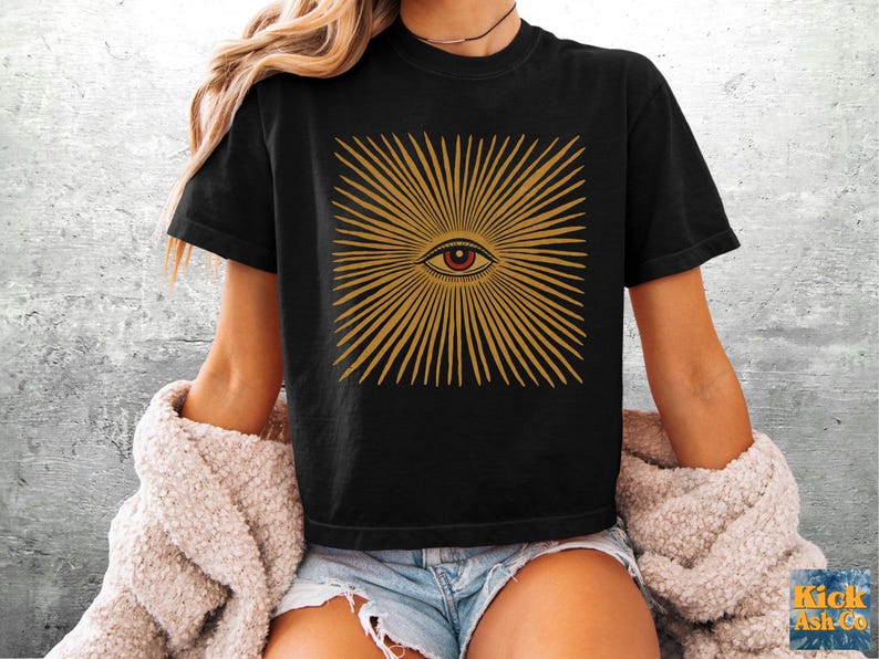 May include: A black short-sleeve t-shirt featuring a graphic print of a central eye surrounded by radiating golden-yellow lines. The eye has a red iris and a black pupil. The design is centered on the chest of the garment.