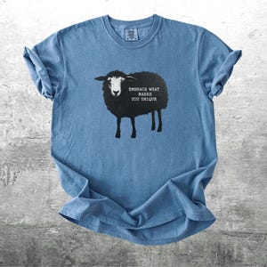 Puede incluir: Camiseta azul con un dise&ntilde;o de oveja negra y el texto "EMBRACE WHAT MAKES YOU UNIQUE". La camiseta tiene mangas enrolladas y cuello redondo. La camiseta es de material suave y se muestra sobre una superficie gris.