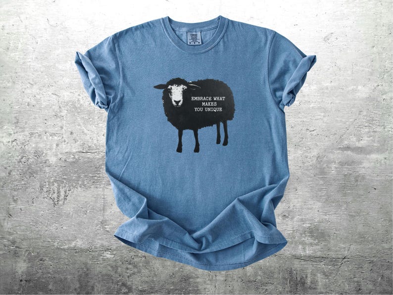 Puede incluir: Camiseta azul con un dise&ntilde;o de oveja negra y el texto "EMBRACE WHAT MAKES YOU UNIQUE". La camiseta tiene mangas enrolladas y cuello redondo. La camiseta es de material suave y se muestra sobre una superficie gris.
