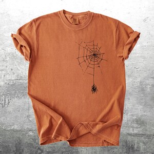 May include: A burnt orange t-shirt featuring a black spider web design with a spider hanging below. The short-sleeved shirt is made of a soft material and is suitable for Halloween or everyday wear.