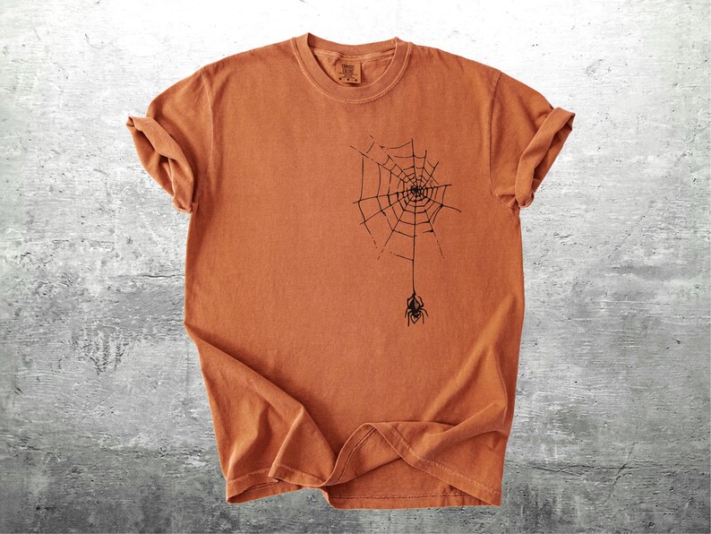 May include: A burnt orange t-shirt featuring a black spider web design with a spider hanging below. The short-sleeved shirt is made of a soft material and is suitable for Halloween or everyday wear.