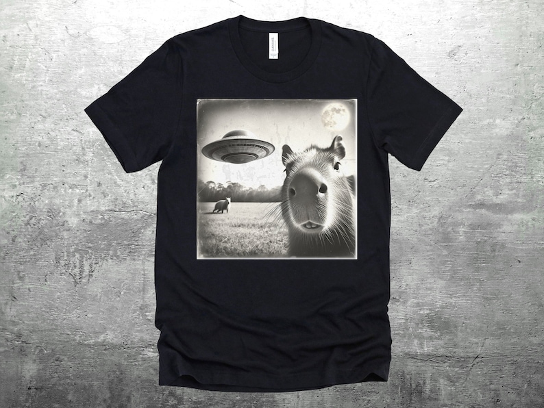K&ouml;nnte beinhalten: Schwarzes T-Shirt mit einem Vintage-Bild eines Capybaras, das in einem Feld zu einem UFO aufblickt. Das Bild ist in Schwarzwei&szlig;.