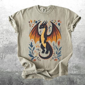 May include: A light gray t-shirt with a black dragon illustration. The dragon has orange and yellow wings and is surrounded by colorful floral designs.