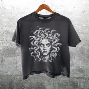 May include: A charcoal gray short-sleeve t-shirt featuring a white graphic print of a face with snakes for hair, resembling Medusa. The design has a distressed, textured appearance. The shirt is displayed on a wooden hanger.