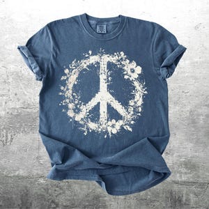 May include: A blue short-sleeved t-shirt featuring a large, distressed white graphic of a peace sign encircled by floral and leaf patterns. The shirt has a crew neckline and a textured, washed-out appearance across the fabric surface.
