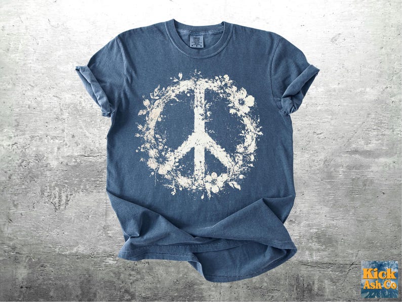 May include: A blue short-sleeved t-shirt featuring a large, distressed white graphic of a peace sign encircled by floral and leaf patterns. The shirt has a crew neckline and a textured, washed-out appearance across the fabric surface.