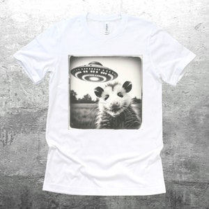 May include: A white t-shirt with a black and white image of an opossum looking up at a flying saucer. The image is in a vintage style frame.