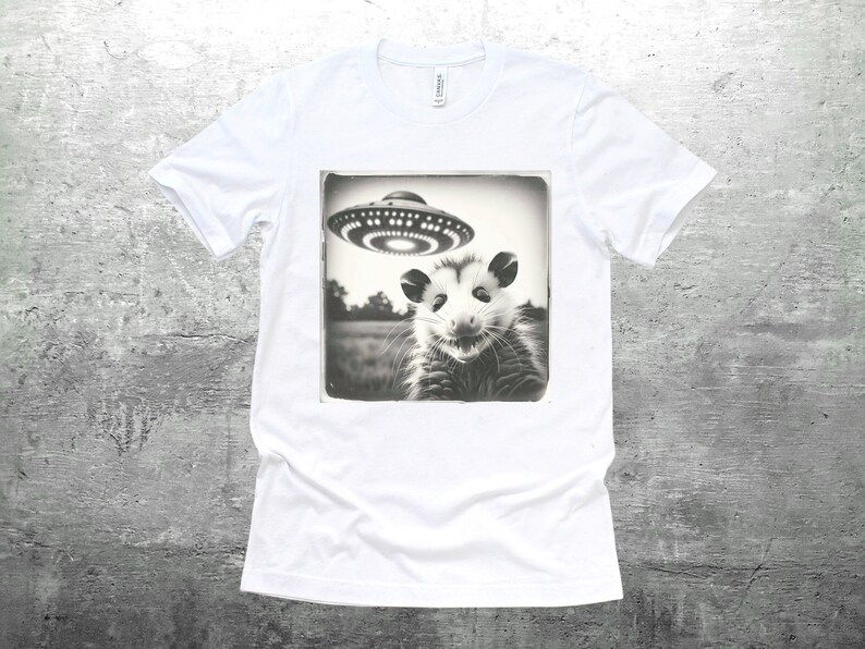 May include: A white t-shirt with a black and white image of an opossum looking up at a flying saucer. The image is in a vintage style frame.
