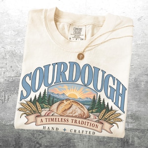 May include: A cream-colored t-shirt featuring a graphic of a loaf of sourdough bread, wheat stalks, pine trees, and a sunrise. The text reads SOURDOUGH, A TIMELESS TRADITION, and HAND CRAFTED. The shirt has a Comfort Colors brand label.