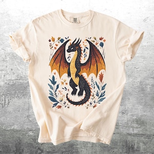 May include: A cream-colored t-shirt with a black dragon illustration. The dragon is surrounded by colorful flowers and leaves.
