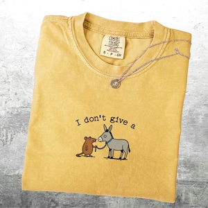 May include: Mustard yellow t-shirt with the phrase "I don't give a" and a cartoon illustration of a mouse and a donkey. A silver necklace with a small pendant rests on the shirt. The shirt is folded.