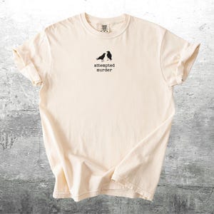 May include: Cream-colored t-shirt with a crew neck. The shirt features a small graphic of two black birds and the text "attempted murder" printed in black below the birds. The shirt is made of soft material.