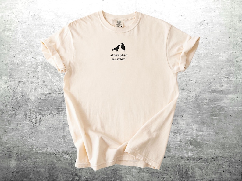 May include: Cream-colored t-shirt with a crew neck. The shirt features a small graphic of two black birds and the text "attempted murder" printed in black below the birds. The shirt is made of soft material.