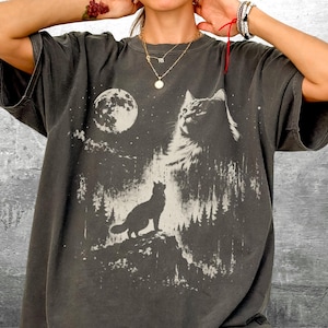 May include: Dark gray t-shirt featuring a graphic of a cat, a full moon, and a forest scene. The cat is silhouetted against the night sky. The shirt has short sleeves and a relaxed fit, suitable for casual wear.