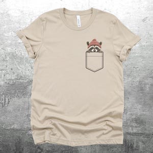 May include: A beige t-shirt with a pocket featuring a raccoon wearing a red beanie hat.