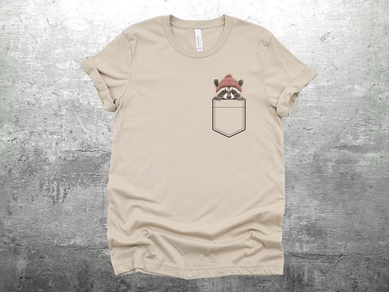 May include: A beige t-shirt with a pocket featuring a raccoon wearing a red beanie hat.