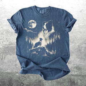 Pu&ograve; includere: T-shirt blu con una grafica di una silhouette di cane su uno sperone roccioso, con una luna piena e una foresta sullo sfondo. Il design &egrave; in stile vintage sbiadito. La maglietta ha le maniche arrotolate.