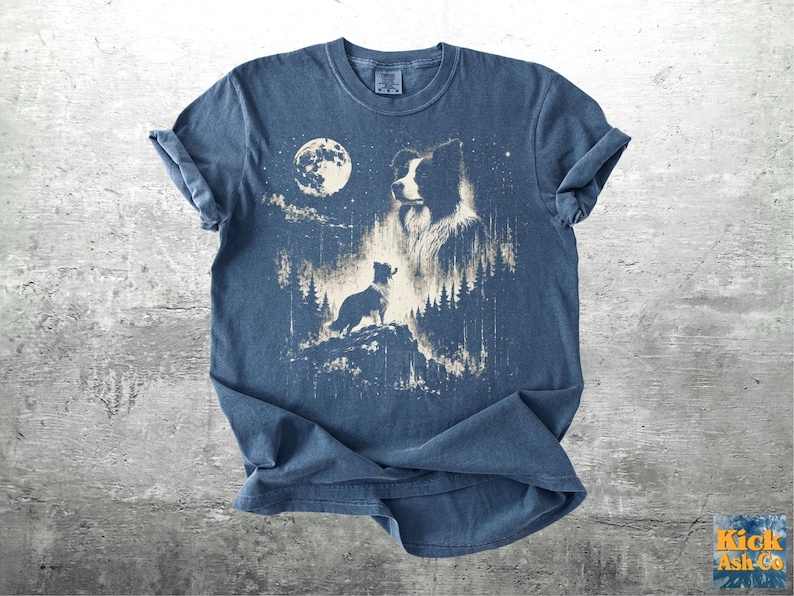Pu&ograve; includere: T-shirt blu con una grafica di una silhouette di cane su uno sperone roccioso, con una luna piena e una foresta sullo sfondo. Il design &egrave; in stile vintage sbiadito. La maglietta ha le maniche arrotolate.