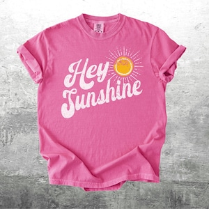 May include: A pink t-shirt with rolled-up sleeves featuring the phrase "Hey Sunshine" in white script, accompanied by a yellow sun graphic. The shirt is made of soft material and is displayed on a gray surface.