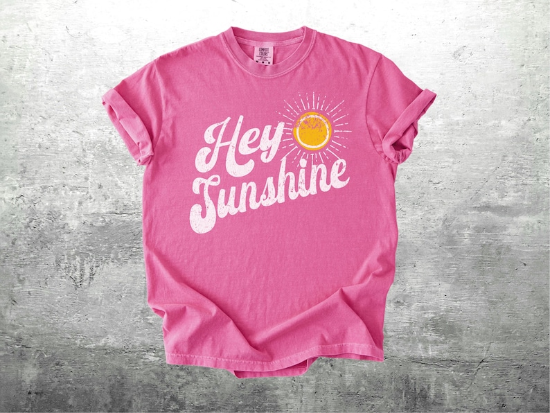 May include: A pink t-shirt with rolled-up sleeves featuring the phrase "Hey Sunshine" in white script, accompanied by a yellow sun graphic. The shirt is made of soft material and is displayed on a gray surface.