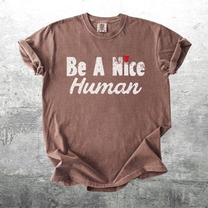 K&ouml;nnte beinhalten: Braunes T-Shirt mit dem Aufdruck "Be A Nice Human" in verblichenen wei&szlig;en Buchstaben. Ein kleines rotes Herz befindet sich &uuml;ber dem Wort "Nice". Das Shirt liegt auf einer grauen, strukturierten Oberfl&auml;che.