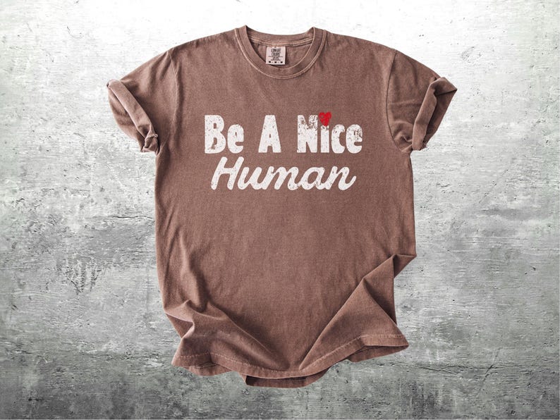 K&ouml;nnte beinhalten: Braunes T-Shirt mit dem Aufdruck "Be A Nice Human" in verblichenen wei&szlig;en Buchstaben. Ein kleines rotes Herz befindet sich &uuml;ber dem Wort "Nice". Das Shirt liegt auf einer grauen, strukturierten Oberfl&auml;che.