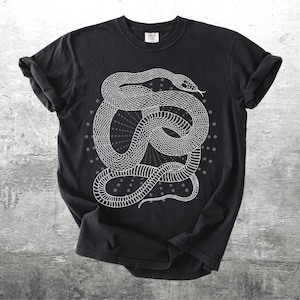 Może przedstawiać: Czarny t-shirt z białą ilustracją zwiniętego węża. Wz&oacute;r węża jest szczeg&oacute;łowy i otoczony wzorem w kropki. T-shirt ma podwinięte rękawy i okrągły dekolt. Koszulka wykonana jest z miękkiego materiału.