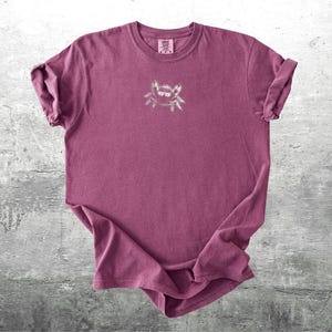 May include: A maroon-colored t-shirt with a small, light-colored graphic on the front. The shirt has rolled-up sleeves and a crew neck. The graphic appears to be a stylized insect or spider.