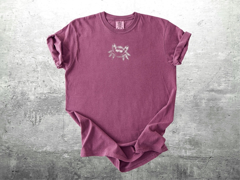 May include: A maroon-colored t-shirt with a small, light-colored graphic on the front. The shirt has rolled-up sleeves and a crew neck. The graphic appears to be a stylized insect or spider.