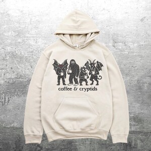 May include: A beige hooded sweatshirt featuring a black graphic of four cryptids holding coffee mugs. The text "coffee & cryptids" is printed below the figures. The sweatshirt has a kangaroo pocket and a drawstring hood.