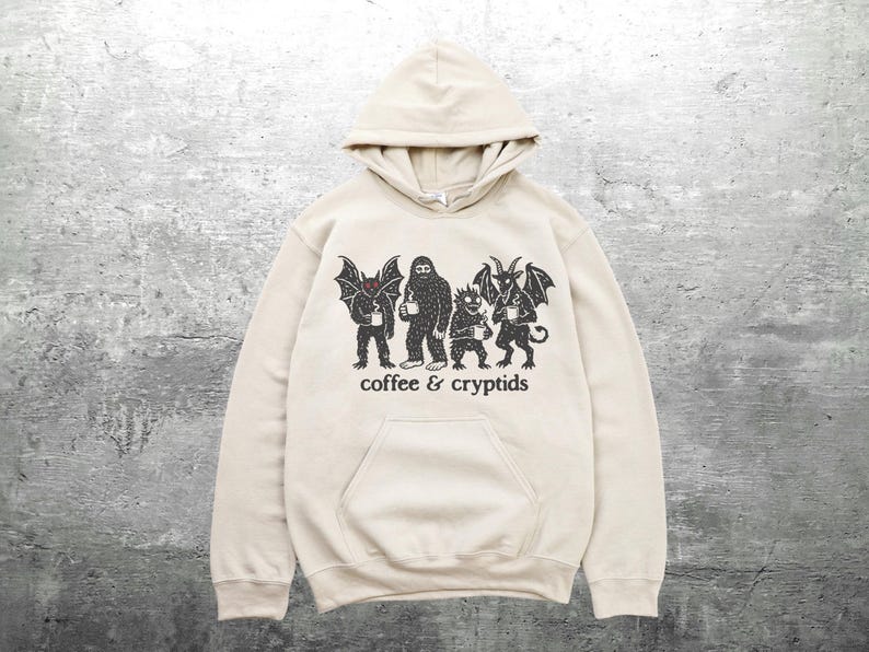 May include: A beige hooded sweatshirt featuring a black graphic of four cryptids holding coffee mugs. The text "coffee & cryptids" is printed below the figures. The sweatshirt has a kangaroo pocket and a drawstring hood.