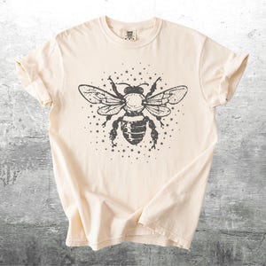 May include: Cream-colored t-shirt featuring a large, detailed illustration of a bee with outstretched wings, surrounded by small stars. The bee design is in dark gray, creating a striking contrast against the shirt's neutral background. The shirt has short sleeves.
