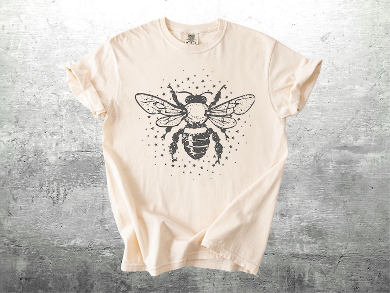 May include: Cream-colored t-shirt featuring a large, detailed illustration of a bee with outstretched wings, surrounded by small stars. The bee design is in dark gray, creating a striking contrast against the shirt's neutral background. The shirt has short sleeves.