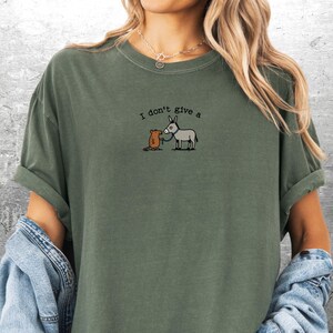 May include: An olive green t-shirt featuring the embroidered text "I don't give a" above a cartoon image of a grey donkey and a brown mouse. The shirt has rolled-up sleeves and is made of a soft material.