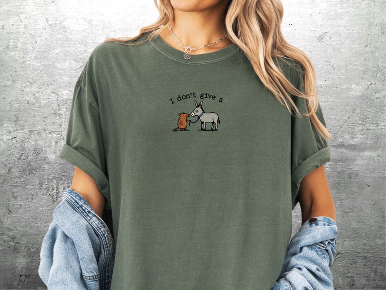 May include: An olive green t-shirt featuring the embroidered text "I don't give a" above a cartoon image of a grey donkey and a brown mouse. The shirt has rolled-up sleeves and is made of a soft material.