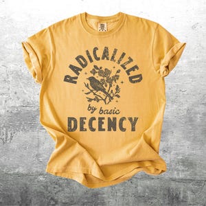May include: Mustard yellow t-shirt with rolled sleeves. The front features a distressed graphic with the words "RADICALIZED" above "by basic DECENCY", and a bird and floral design. The shirt is laid flat on a gray surface.