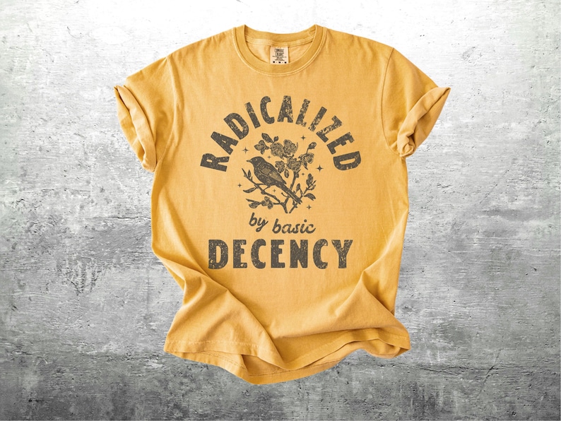 May include: Mustard yellow t-shirt with rolled sleeves. The front features a distressed graphic with the words "RADICALIZED" above "by basic DECENCY", and a bird and floral design. The shirt is laid flat on a gray surface.