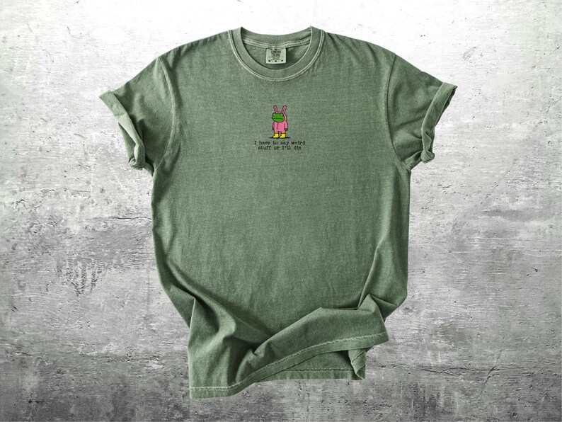 May include: An olive green t-shirt featuring a small graphic of a frog in a bunny costume. The text "I have to say weird stuff or I'm me" is printed below the graphic. The sleeves are rolled up.