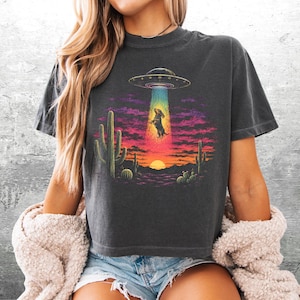Puede incluir: Una camiseta gris carb&oacute;n con un estampado gr&aacute;fico de un perro salchicha siendo abducido por un OVNI sobre un paisaje des&eacute;rtico con cactus y una puesta de sol. El dise&ntilde;o incluye colores vibrantes como amarillo, naranja, rosa y azul en el haz de luz.