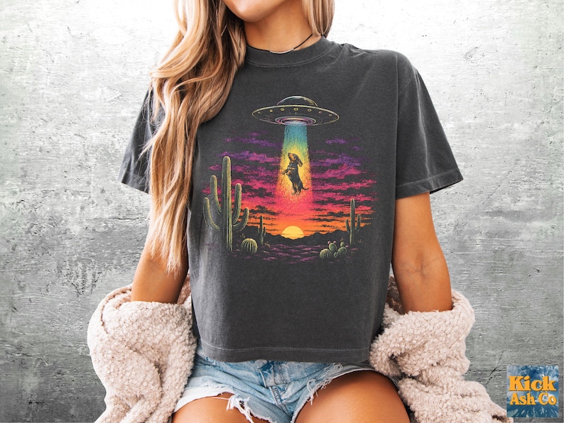 Puede incluir: Una camiseta gris carb&oacute;n con un estampado gr&aacute;fico de un perro salchicha siendo abducido por un OVNI sobre un paisaje des&eacute;rtico con cactus y una puesta de sol. El dise&ntilde;o incluye colores vibrantes como amarillo, naranja, rosa y azul en el haz de luz.