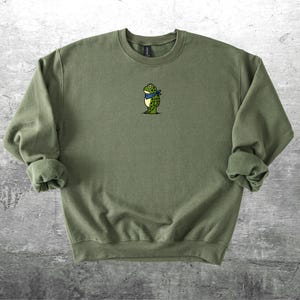 Puede incluir: Sudadera verde oliva con cuello redondo y mangas largas. Un peque&ntilde;o bordado de una rana de dibujos animados con un pa&ntilde;uelo azul est&aacute; centrado en el pecho. La sudadera es de material suave.