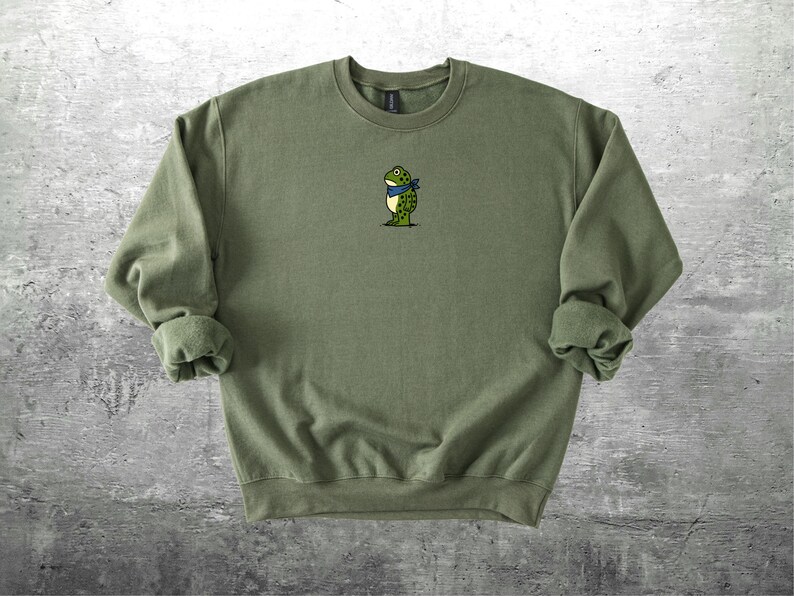 Puede incluir: Sudadera verde oliva con cuello redondo y mangas largas. Un peque&ntilde;o bordado de una rana de dibujos animados con un pa&ntilde;uelo azul est&aacute; centrado en el pecho. La sudadera es de material suave.