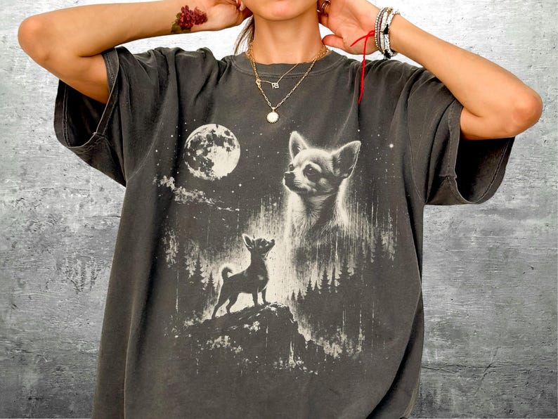 May include: Dark gray t-shirt featuring a detailed graphic of a Chihuahua dog under a full moon and starry night sky. The design includes a silhouette of a dog on a hill and a portrait of a dog's face. The shirt is a casual, oversized fit.