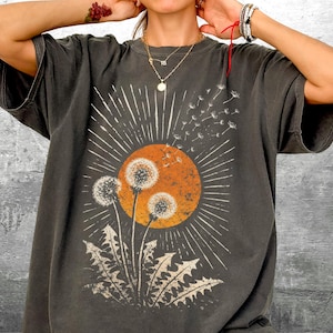 May include: Dark gray t-shirt featuring a dandelion design with an orange sun and white rays. The graphic tee has a vintage, distressed look, with a crew neck and short sleeves. The dandelion graphic is in the center of the shirt.