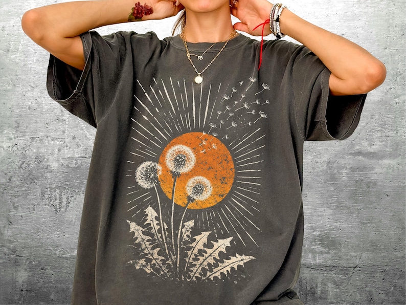 May include: Dark gray t-shirt featuring a dandelion design with an orange sun and white rays. The graphic tee has a vintage, distressed look, with a crew neck and short sleeves. The dandelion graphic is in the center of the shirt.