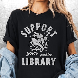 May include: Black t-shirt with the text "SUPPORT your public LIBRARY" in distressed white lettering. A bird perched on a flowering branch is centered above the text. The shirt has rolled-up sleeves.
